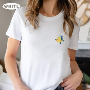 May include: White t-shirt with a small, embroidered cartoon fish in yellow, blue, and black. The shirt has short sleeves and a crew neck. The word "WHITE" is in a white oval in the upper left corner.