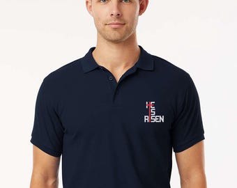 May include: A navy blue polo shirt with short sleeves. The shirt has the text "HE IS RISEN" in a vertical design on the left chest. The text is white with red accents.