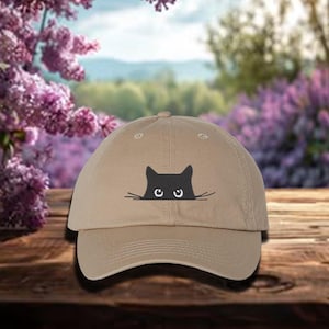 May include: A tan baseball cap with a black cat face peeking out from behind the brim. The cat has large, round eyes and a small, black nose.