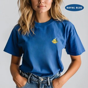 May include: Royal blue t-shirt with a small, embroidered yellow and blue cartoon fish on the chest. The shirt has short sleeves and a crew neck. The text "ROYAL BLUE" is in a blue oval in the upper right corner.