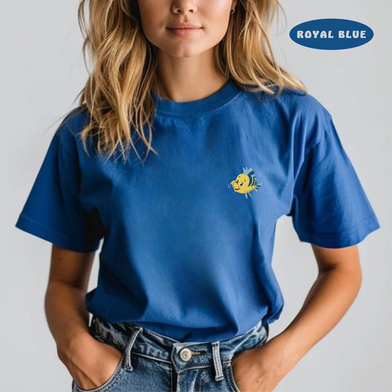 May include: Royal blue t-shirt with a small, embroidered yellow and blue cartoon fish on the chest. The shirt has short sleeves and a crew neck. The text "ROYAL BLUE" is in a blue oval in the upper right corner.
