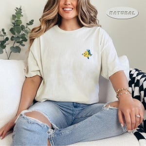 May include: Cream-colored t-shirt with a small, embroidered yellow and blue fish design on the chest. The word "NATURAL" is printed on the top right of the shirt. The shirt is paired with light blue jeans.
