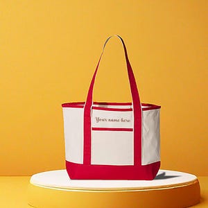 May include: A white tote bag with red accents, including the base, handles, and trim. The bag features a front pocket with the text "Your name here" in a script font. The bag is displayed on a white platform against a yellow background.