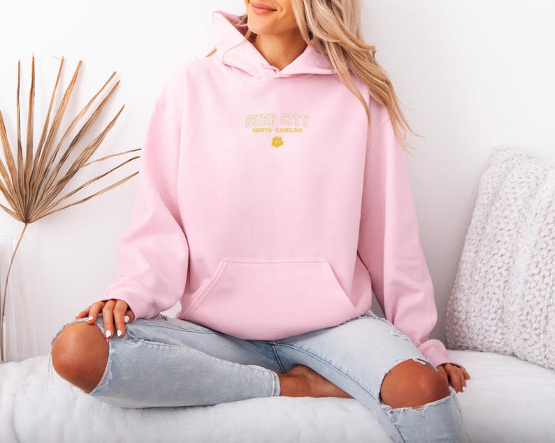 May include: A light pink hoodie with a front pocket and a hood. The text "Surf City North Carolina" is printed in a gold color on the front. A small yellow flower graphic is below the text. The person is wearing light blue jeans.