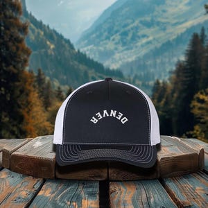 May include: A black and white trucker hat with the word "DENVER" arched across the front in white lettering. The hat has a black front panel and a white mesh back. The hat is sitting on a wooden surface with a mountain backdrop.