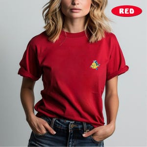 May include: Red t-shirt with a small, embroidered cartoon fish on the chest. The shirt has short sleeves and is made of a soft material. The word "RED" is in a red oval in the top right corner.
