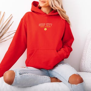 May include: A red hoodie with the text "SURF CITY NORTH CAROLINA" in yellow lettering. The hoodie has a front pocket and a drawstring hood. The model is wearing ripped blue jeans.