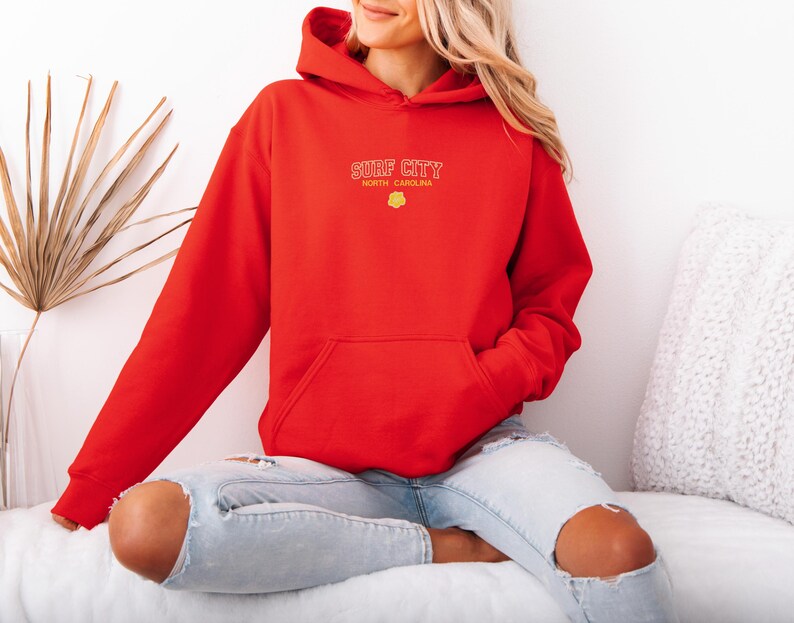 May include: A red hoodie with the text "SURF CITY NORTH CAROLINA" in yellow lettering. The hoodie has a front pocket and a drawstring hood. The model is wearing ripped blue jeans.