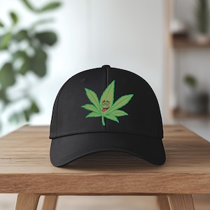 May include: Black baseball cap featuring a green cannabis leaf with a smiling face and a red tongue. The cap is displayed on a wooden surface. The cannabis leaf design is centered on the front of the cap.
