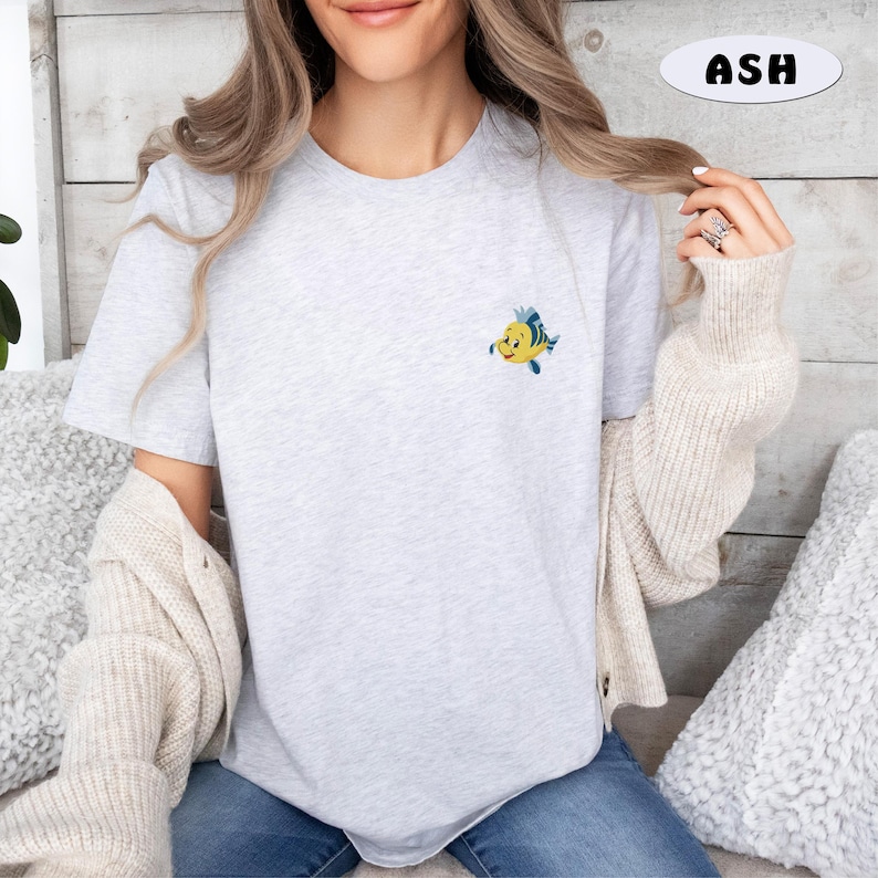 May include: A light gray t-shirt with a small, embroidered cartoon fish in yellow, blue, and white. The word "ASH" is in a white oval in the upper right corner. The shirt is short-sleeved.