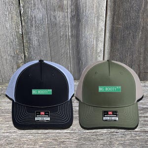 May include: Two trucker hats with the text "BIG BOOTY BLVD" on a green street sign. One hat is black with a white mesh back, the other is olive green with a tan mesh back. Both have a Richardson 112 tag.