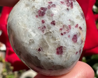 May include: A smooth, rounded stone with a white and gray base color, speckled with small, vibrant, ruby-red crystals. The polished surface of the stone reflects light, highlighting the contrasting colors and textures. This natural stone is a unique decorative item.