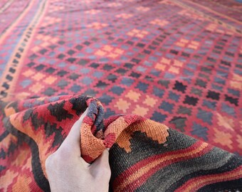 May include: A close-up of a red, orange, and black patterned rug. The rug is folded in half and a hand is holding the fold.