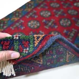 May include: A section of a red, hand-knotted rug with a detailed geometric pattern. The rug's border features a blue and green design, with a fringe at the end. The rug is being held up, revealing its intricate weave and colour variations.