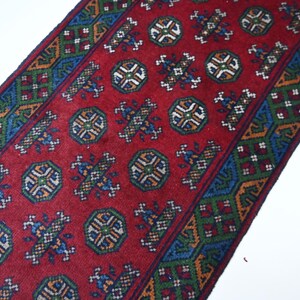 Puede incluir: Una alfombra roja con un patrón geométrico repetitivo. La alfombra presenta un campo central rojo con medallones repetidos y un borde de diseños geométricos verdes, azules y dorados. La alfombra está en ángulo diagonal.