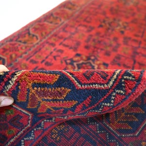 May include: Close-up of a red oriental rug with intricate geometric patterns. The rug features a deep red field with navy blue and gold accents in the border and design. The rug is folded over, revealing the detailed weave.