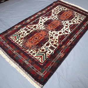 Puede incluir: Una alfombra persa tradicional con fondo blanco y intrincados patrones en rojo, azul y marrón. La alfombra tiene un borde con flecos.