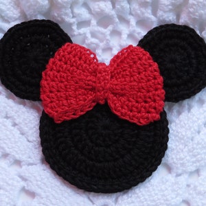 May include: Crocheted black mouse ears with a red bow. The ears are circular and the bow is centered between them. The crochet work is visible, with a textured appearance. The design is set against a white, textured background.