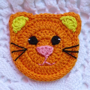 May include: A crocheted orange cat face with yellow ears, black eyes, a pink nose, and black whiskers. The handmade cat face is likely a patch or applique for crafts, with a textured appearance.