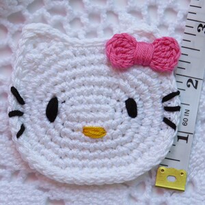 Puede incluir: Una cara de Hello Kitty blanca tejida a ganchillo con ojos y bigotes negros, una nariz amarilla y un lazo rosa. El art&iacute;culo mide aproximadamente 5 cm de di&aacute;metro. El fondo es una superficie blanca tejida a ganchillo.