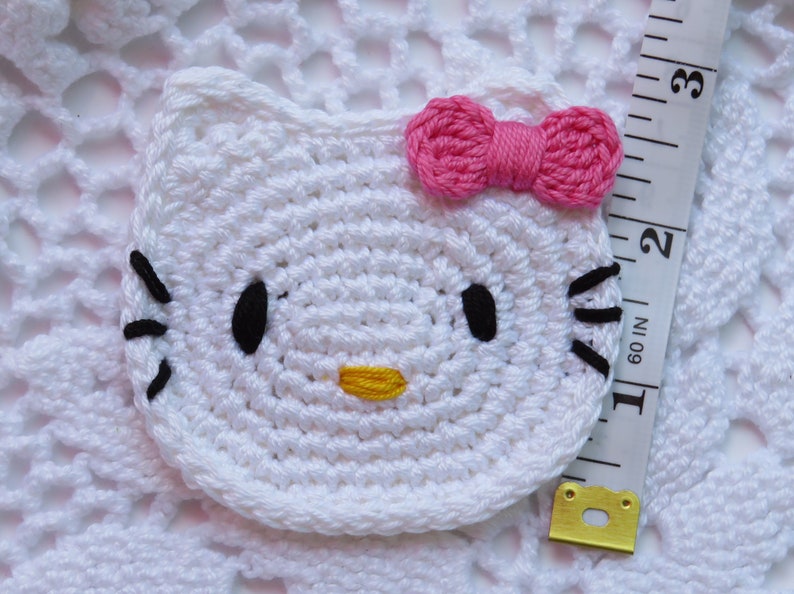 Puede incluir: Una cara de Hello Kitty blanca tejida a ganchillo con ojos y bigotes negros, una nariz amarilla y un lazo rosa. El art&iacute;culo mide aproximadamente 5 cm de di&aacute;metro. El fondo es una superficie blanca tejida a ganchillo.
