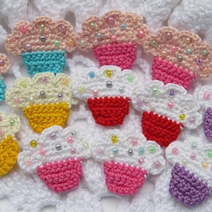 May include: A collection of colorful, crocheted cupcake-shaped appliques. Each cupcake features a different color frosting and base, with beaded sprinkles. The appliques are arranged on a white, textured surface.