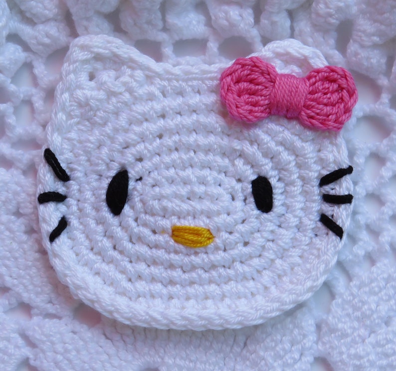 Puede incluir: Una cara de gato blanca tejida a ganchillo con ojos y bigotes negros, una nariz amarilla y un lazo rosa. El art&iacute;culo hecho a mano es probablemente un posavasos o una aplicaci&oacute;n, que muestra un personaje de dibujos animados popular.