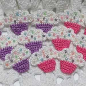 May include: Crocheted cupcake appliques with white frosting, purple and pink cupcake liners, and colorful bead sprinkles. These handmade appliques are perfect for embellishing crafts and adding a whimsical touch to projects.