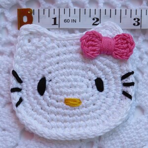 Puede incluir: Una cara de Hello Kitty blanca tejida a ganchillo con ojos y bigotes negros, una nariz amarilla y un lazo rosa. El art&iacute;culo tejido a ganchillo mide aproximadamente 6,35 cm de ancho. El art&iacute;culo est&aacute; colocado sobre una superficie blanca de tejido abierto.