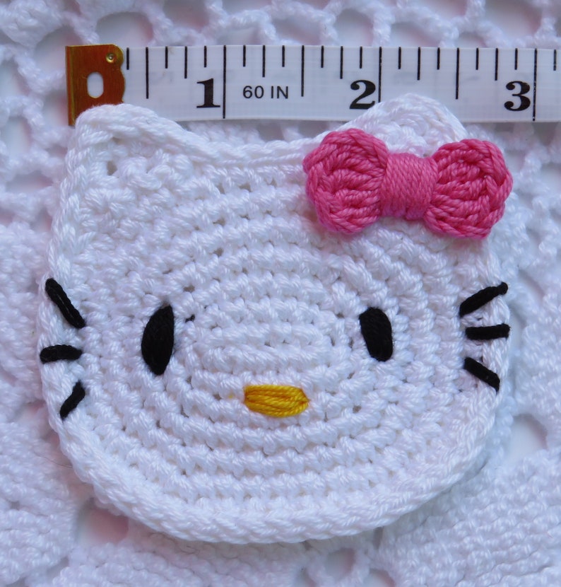 Puede incluir: Una cara de Hello Kitty blanca tejida a ganchillo con ojos y bigotes negros, una nariz amarilla y un lazo rosa. El art&iacute;culo tejido a ganchillo mide aproximadamente 6,35 cm de ancho. El art&iacute;culo est&aacute; colocado sobre una superficie blanca de tejido abierto.