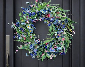 Peut inclure: Une couronne florale vibrante avec des fleurs bleues, roses et blanches, agrémentée d'un feuillage vert. La couronne est affichée sur une porte gris foncé. Le texte en bas indique "blue flower".