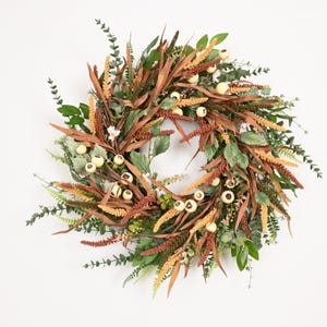 Pode incluir: Uma guirlanda circular com uma mistura de folhagem artificial, folhas longas castanhas com aspeto seco, espigas florais texturizadas em laranja e amarelo, e pequenas bagas redondas de cor creme. O arranjo é denso e composto por várias folhagens.