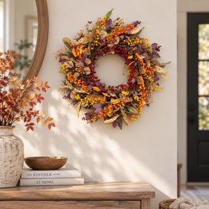 May include: A circular autumn wreath featuring a dense arrangement of faux foliage, including yellow, orange, and deep red berries, along with variegated leaves in shades of gold, brown, and muted purple, all secured to a natural twig base structure.