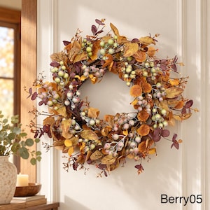 Puede incluir: Una corona circular que presenta una mezcla de follaje de colores otoñales en tonos dorados, marrones y morado oscuro, intercalados con racimos de bayas de color verde pálido y rosa claro. El texto Berry05 está impreso en la esquina inferior derecha.