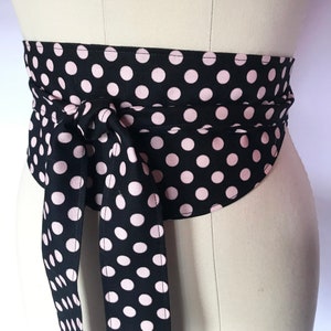 May include: A black fabric wrap belt with a pink polka dot pattern. The belt is tied in a bow at the front, with long ties hanging down. The belt is wrapped around a mannequin.