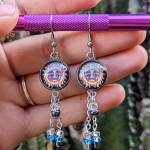 May include: A pair of silver dangle earrings with a sun face charm and blue and clear beads.