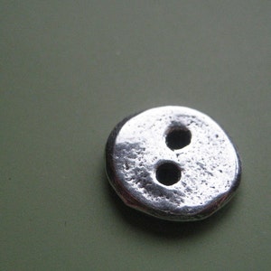 May include: A close-up of a silver-colored button with two holes. The button has a textured surface and a rounded shape. The button is likely made of metal and is set against a green background.
