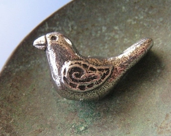 May include: A small, silver-colored bird figurine with detailed wing and body patterns. The bird is perched on a textured, bronze-colored surface, creating a contrast in color and texture. The figurine's design is simple yet elegant.