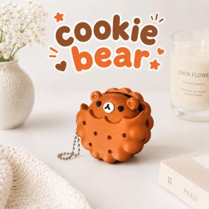 以下が含まれることがあります： 中央から小さなクマの顔がのぞく、クッキーの形をした茶色のワイヤレスイヤホン用保護ケース。ケースの表面は質感のある仕上がりで小さな凹みがあり、シルバーカラーの金属製ボールチェーンが付いています。