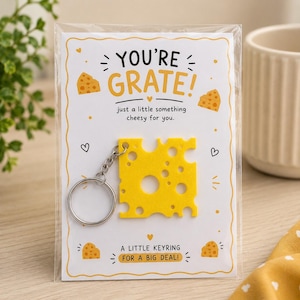 Peut inclure: Un porte-clés jaune en forme de fromage fixé sur une carte de présentation blanche. La carte porte les inscriptions "YOU'RE GRATE!", "just a little something cheesy for you." et "A LITTLE KEYRING FOR A BIG DEAL!" avec des illustrations.