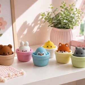 Può includere: Una collezione di sei piccole statuette di animali, ognuna inserita in un vasetto colorato in miniatura. Il set comprende un orso marrone, un coniglio bianco, un pinguino blu, un pulcino giallo, una volpe arancione e un ippopotamo grigio.