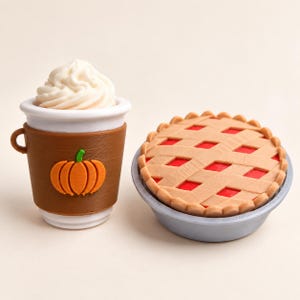 Pode incluir: Uma chávena miniatura branca com uma manga castanha com um gráfico de abóbora laranja, coberta com natas batidas brancas, ao lado de uma tarte miniatura redonda com crosta de rede bege e recheio vermelho num prato circular cinzento.