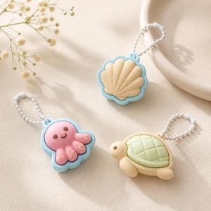 Peut inclure: Trois porte-clés sur le thème marin : un coquillage, une pieuvre et une tortue. Le coquillage est beige avec un bord bleu clair. La pieuvre est rose avec un bord bleu clair. La tortue est beige avec une carapace verte. Chaque porte-clés a une chaîne argentée.