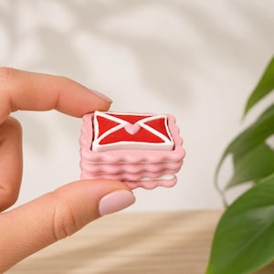 May include: A miniature, pink, layered decorative item, like a stack of biscuits, with a red and white envelope design on top. The envelope features a heart. The item is held between fingers against a soft background.