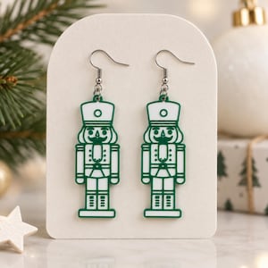 Può includere: Un paio di orecchini pendenti con una figura di schiaccianoci bianca e piatta con un contorno verde scuro. Il design dello schiaccianoci include un cappello alto, una giacca con bottoni e stivali. Gli orecchini pendono da ganci in metallo argentato.
