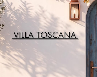 May include: A black metal wall sign featuring the text VILLA TOSCANA in a clean sans-serif font. The sign is mounted on a smooth off-white exterior wall next to an arched blue wooden door, a copper wall lantern, and potted flowering plants.
