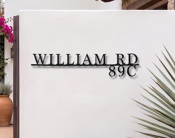 May include: A black metal address sign mounted on a white wall, featuring the text "WILLIAM RD" on the top line and "89C" on the bottom line. The sign is constructed from thin, dark, cut-out lettering that stands out against the smooth surface.