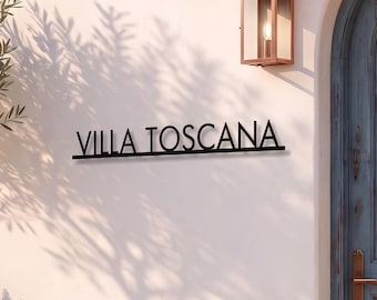 May include: A black metal sign with the text VILLA TOSCANA mounted on a white wall next to an arched blue wooden door. A copper lantern hangs above the door, which is framed by orange bougainvillea flowers and potted lavender plants.