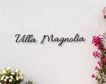 May include: A white exterior wall features black metal script lettering that reads Villa Magnolia. To the right, a wooden door sits beneath a tiled roof overhang. Three terracotta pots contain white flowers, yellow flowers, and succulents.