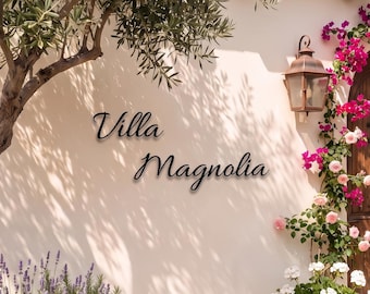 May include: A beige stucco wall features the black text Villa Magnolia in a cursive font. An arched wooden door is framed by vibrant pink bougainvillea and white flowers in a terracotta pot. An olive tree and lavender plants are visible.